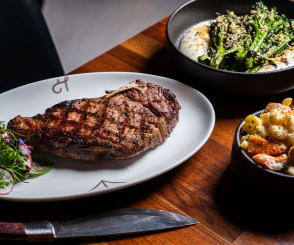 a large steak on a plate with potatoes and broccoli as sides at Huntress